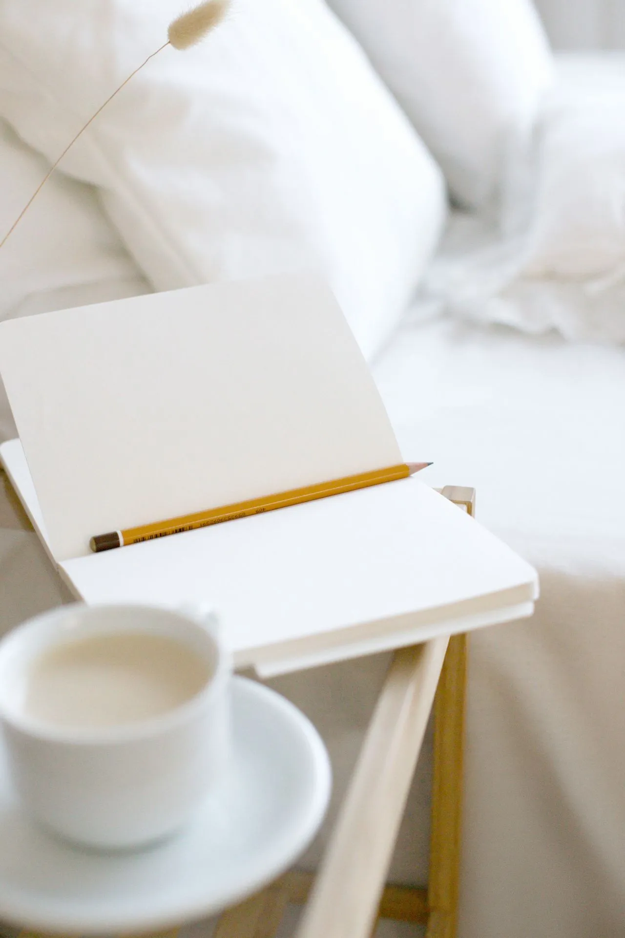 Open notebook, a pencil and a cup of tea on a bedside table beside white bedding
