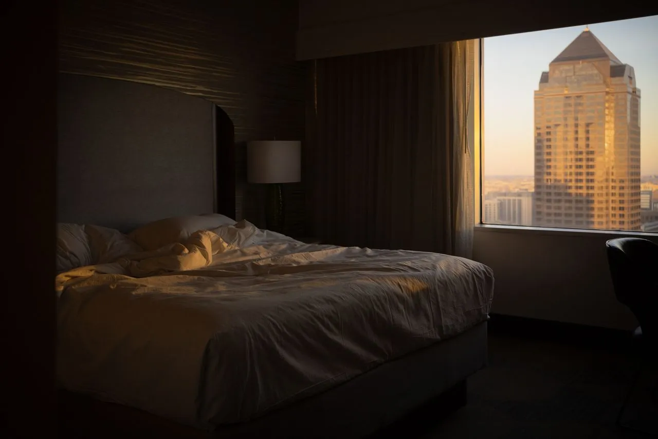 Dim hotel bedroom with a made bed beside a window onto a tall tower at dusk