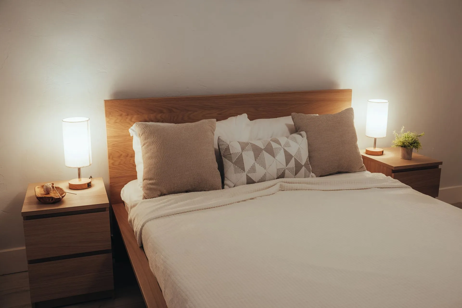 Neatly made bed with beige cushions between two softly glowing bedside lamps at night