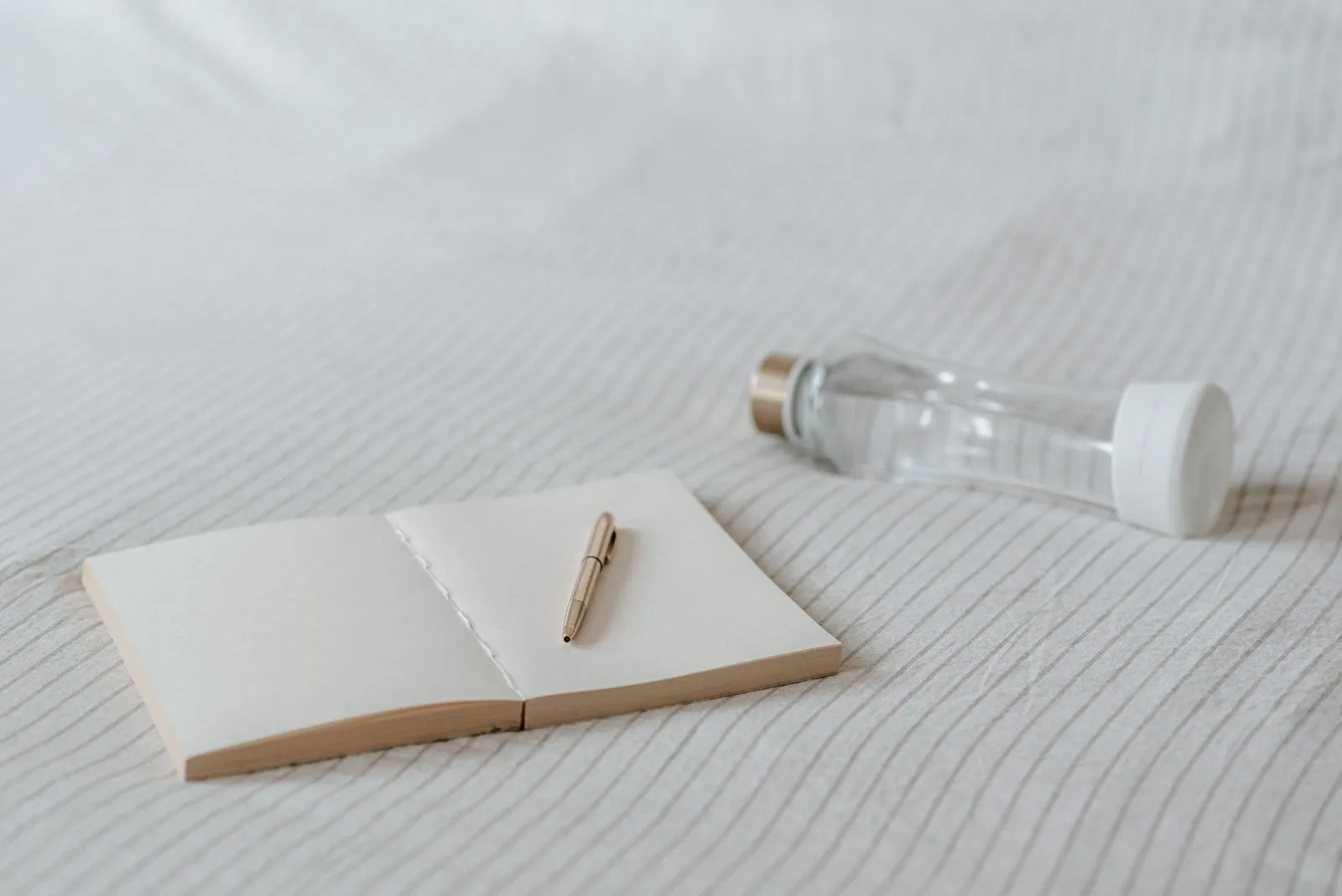 Open blank notebook with a gold pen and a water bottle on bedding