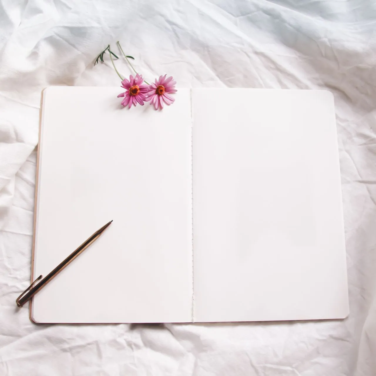 Blank open notebook and a slim pen on crumpled white bedding, two small pink flowers at the top