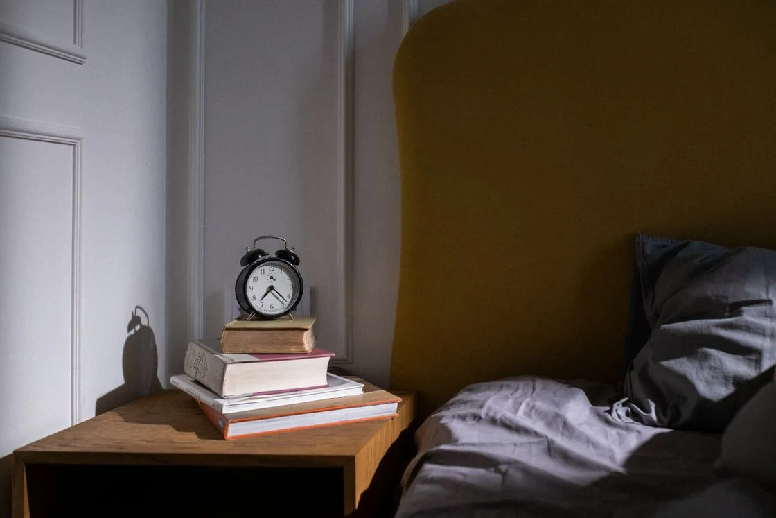 Plain black alarm clock on a stack of books on a wooden nightstand beside a made bed